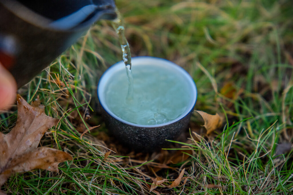 A ceramic cup sits in the grass, while Emerald Leaf Organic Baozhong is poured into it